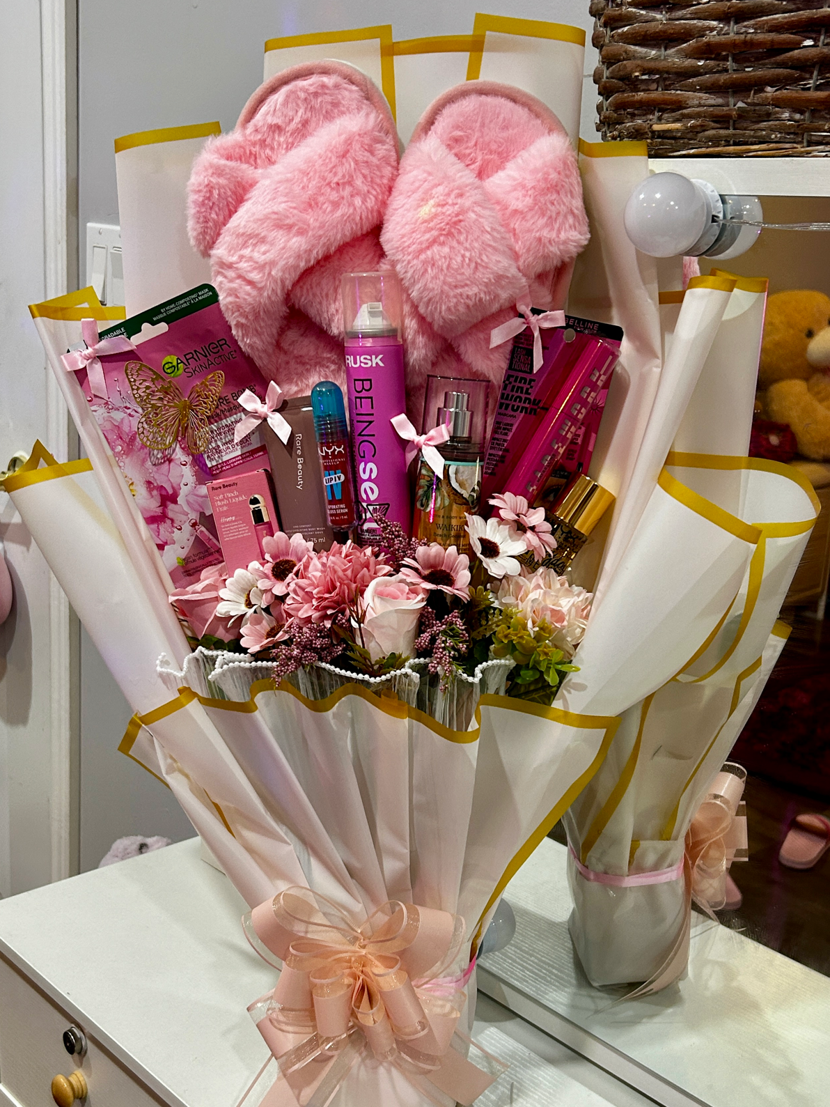 Cosmetic gift bouquet with beauty products, pink slippers, and flowers wrapped in white paper with a pink bow.